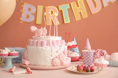 Koyu bej duvarın yanındaki beyaz masada mumlar ve şekerler olan doğum günü pastası.