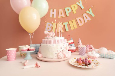 Koyu bej duvarın yanındaki beyaz masada mumlar ve şekerler olan doğum günü pastası.