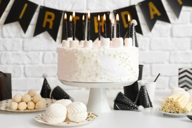 Doğum günü pastası, beyaz masada mumlar ve tuğla duvarda çelenkler.