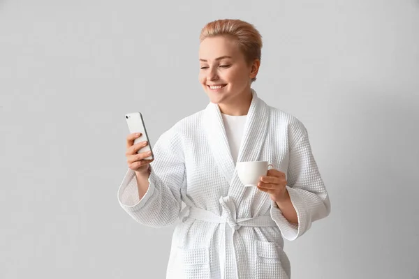 Młoda Kobieta Szlafroku Filiżanką Kawy Telefonem Komórkowym Jasnym Tle — Zdjęcie stockowe