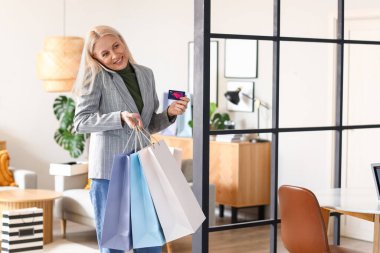 Kredi kartı ve alışveriş torbalarıyla evde cep telefonuyla konuşan olgun bir kadın.
