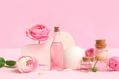 Gül özlü kozmetik yağı şişeleri ve pembe arka planda çiçekler.