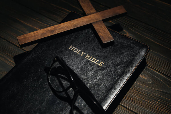 Holy Bible, cross and eyeglasses on wooden background, closeup