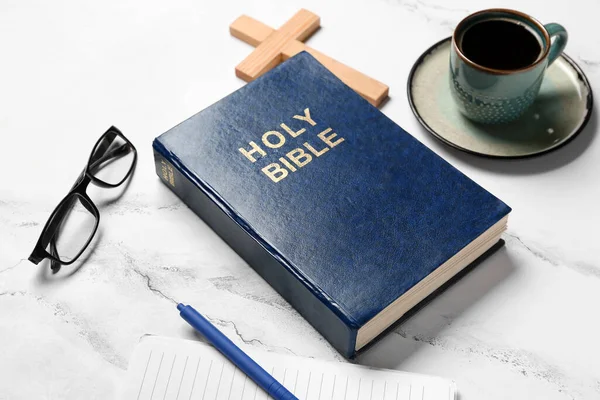 Holy Bible, eyeglasses and cup of coffee on white marble background