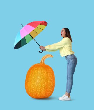 Şok olmuş genç bir kadın gökkuşağı şemsiyesi ve mavi arka planda büyük bir balkabağı