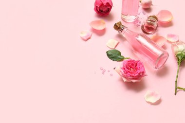 Gül özlü kozmetik yağı şişeleri ve pembe arka planda çiçekler.