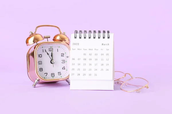 紫色背景上装有闹钟和眼镜的三月折页纸日历 — 图库照片