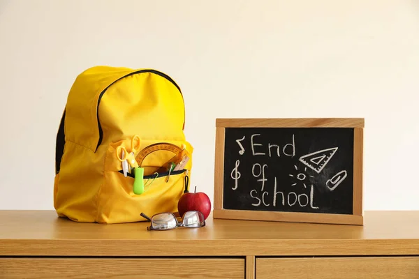 stock image Chalkboard with text END OF SCHOOL, apple and backpack on table near light wall