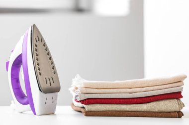 Elektrikli ütü ve üst üste dizilmiş giysiler beyaz masada.