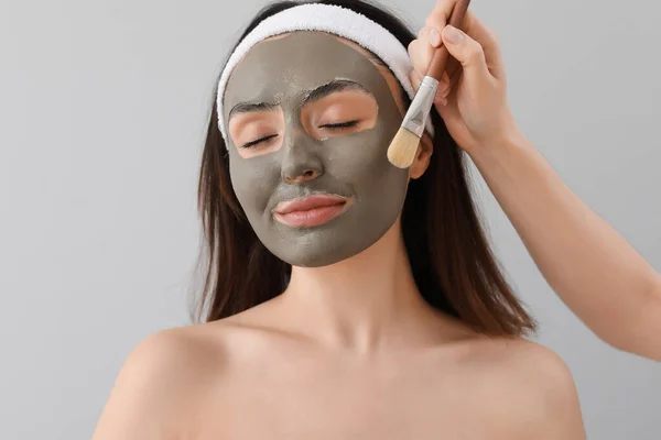 Cosmetoloog Aanbrengen Van Klei Masker Gezicht Jonge Vrouw Grijze Achtergrond — Stockfoto