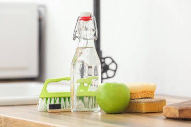 Bottle of vinegar, brushes, sponges and apple on wooden table clipart