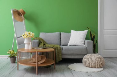 Yeşil duvarın yanında gri kanepesi, aynası ve sehpası olan şık bir oturma odası.