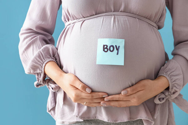 Young pregnant woman and paper with word BOY on her belly against blue background, closeup