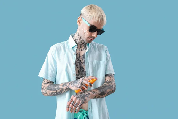 Tattooed man applying sunscreen cream on light blue background