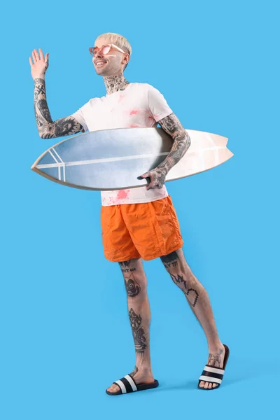 Młody Wytatuowany Mężczyzna Deską Surfingową Machającą Ręką Jasnoniebieskim Tle — Zdjęcie stockowe