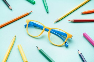 Soluk turkuaz arka planda renkli kalemlerle süslü sarı gözlükler.