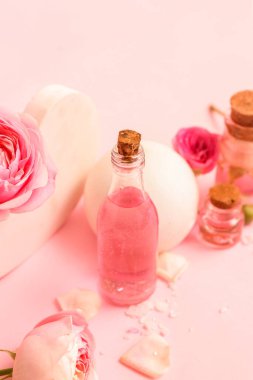 Gül özlü kozmetik yağı şişeleri ve pembe arka planda çiçekler.