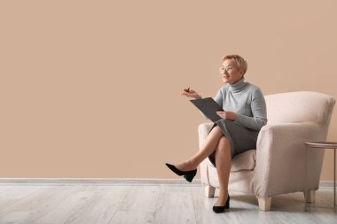 Olgun psikolog bej duvarın yanındaki koltukta pano ile çalışıyor.