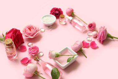Gül özlü kozmetik yağı şişeleri ve pembe arka planda çiçekler.