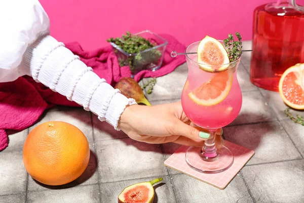 ピンクの壁の近くのタイルテーブルの上にイチジク グレープフルーツとタイムと新鮮な夏のカクテルのガラスを保持女性 — ストック写真