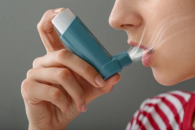 Gri arka planda astım spreyi kullanan genç kadın, yakın plan.