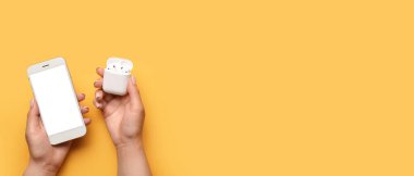 Sarı arka planda kablosuz kulaklıklı modern cep telefonlu, metin için yeri olan kadın eller