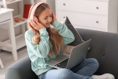 Kulaklıklı, dizüstü bilgisayarlı küçük bir kız evdeki kanepede müzik dinliyor.