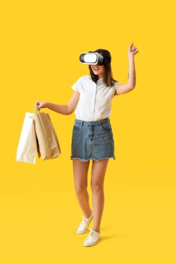 Sarı arka planda alışveriş torbaları olan sanal gerçeklik gözlüklü güzel bir kadın.