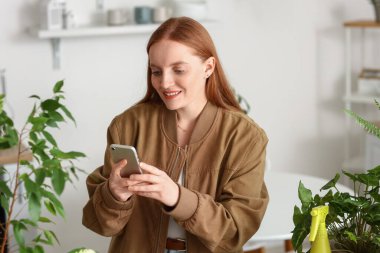Young woman with green houseplants using mobile phone at home
