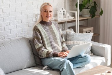 Evde kanepede dizüstü bilgisayarla çalışan olgun bir kadın programcı.