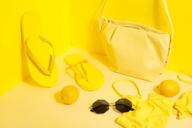 Sarı arka planda mayo, güneş gözlüğü ve çantalı terlikler.