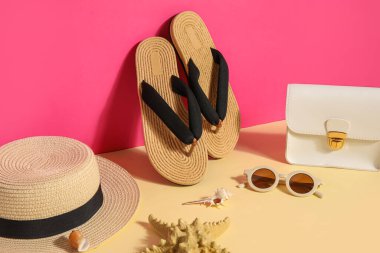 Güneş gözlüklü, çantalı ve hasır şapkalı terlikler pembe duvarın yanında.