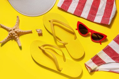 Sarı arka planda mayo, güneş gözlüğü ve tenis şapkası olan parmak arası terlikler.