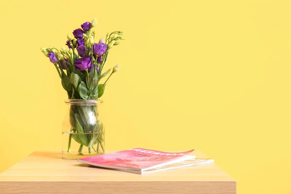 Vase with eustoma flowers and magazine on table near yellow wall