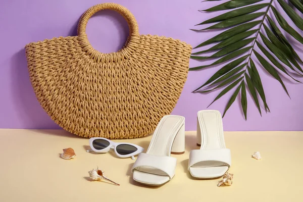 Pair of high heeled sandals with sunglasses and wicker bag near lilac wall