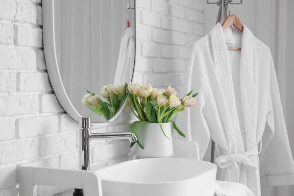 stock image Mirror hanging on white brick wall near sink, vase with tulip flowers and bathrobe