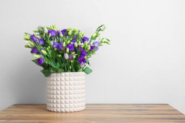 Beyaz duvarın yanındaki ahşap masada, östoma çiçekleri olan vazo.