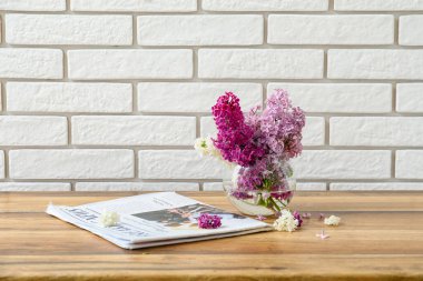 Beyaz tuğla duvarın yanındaki ahşap masada güzel kokulu leylak çiçekleri ve dergi vazosu.