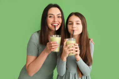 Annesiyle birlikte yeşil arka planda smoothie içen küçük kız.