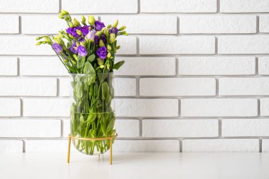 Beyaz tuğla duvarın yanındaki masada, östoma çiçekleri olan vazo.