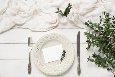 Çatal bıçak takımı, boş kart ve beyaz ahşap arka planda bitki dalları.