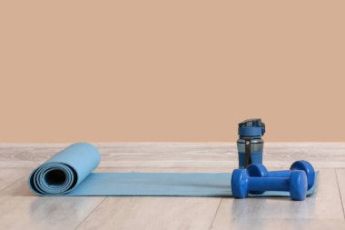 Spor salonundaki bej duvarın yanında halterli ve şişeli yoga minderi.