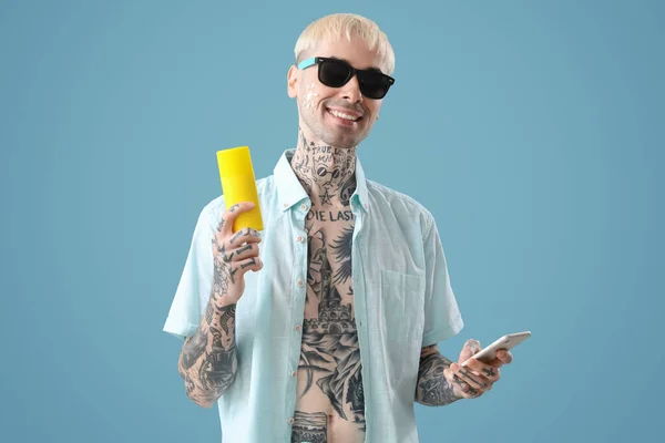 Tattooed man with phone and sunscreen cream on light blue background