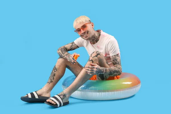 Tattooed man with sunscreen cream and swimming ring on light blue background