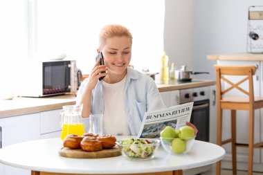 Gazeteli genç bir kadın mutfakta cep telefonuyla konuşuyor.