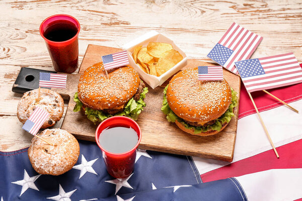 Board with tasty burgers, potato chips, muffins, cola and American flag on light wooden background. Memorial Day celebration
