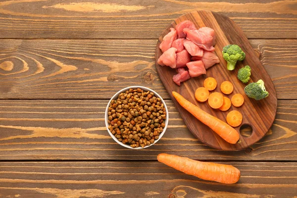 stock image Bowl with dry pet food, raw meat and natural products on wooden background