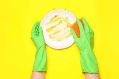 Lastik eldivenli kadın elleri sarı arka planda kirli tabakları yıkıyor.