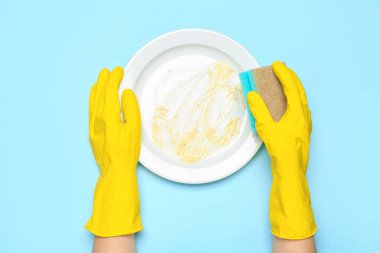 Lastik eldivenli kadın elleri mavi arka planda kirli tabakları yıkıyor.