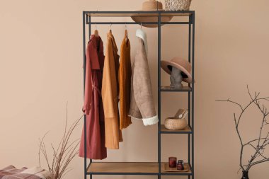 Salondaki bej duvarın yanında elbise ve şapkalı raf ünitesi.